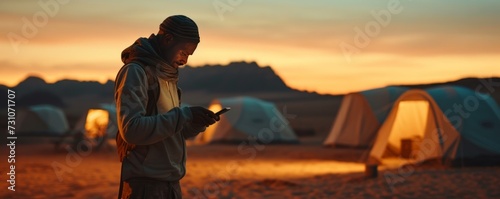 In a medium shot, a man gazes at his smartphone while at a desert camp during sunset, blending modern technology with the timeless beauty of the desert landscape.