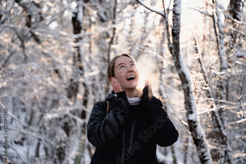 Portrait of young cheerful woman in outdoor gear looking up and admiring nature while out hiking in the winter forest, a tourist stands in a magical snowy frosty wood and admires the beauty of nature