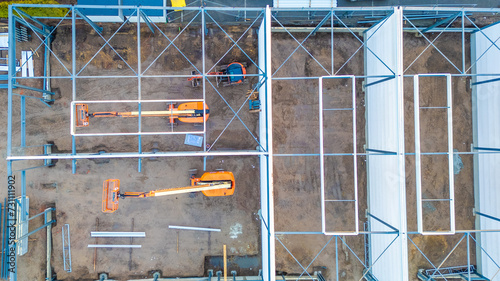 This aerial image provides a bird s-eye view of a construction site  showcasing the geometric arrangement of scaffolding and machinery. Two orange telescopic boom lifts stand out against the muddy