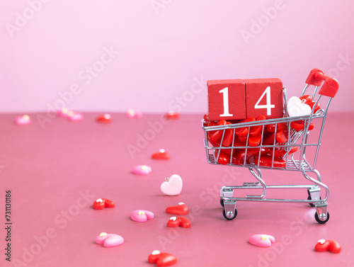 A red wood cube calendar with number 14 in a mini shopping cart with small red heart lying in it, isolated pink background. Valentine day concept 