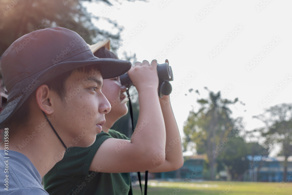 Obraz premium Asian teenboys learning nature by using binoculars to watch birds and insects in public park during summer camp of their school, idea for learning creatures and wildlife animals outside the classroom.