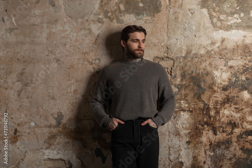 Handsome brutal man with a beard and hairstyle in a fashion knitted sweater and black jeans near a concrete grunge wall