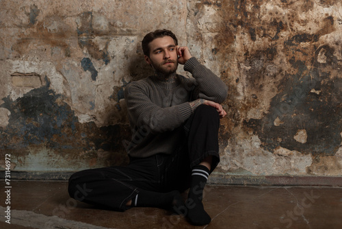 Cool hipster man in a knitted sweater and black jeans with socks sits near an old grunge concrete wall in the studio
