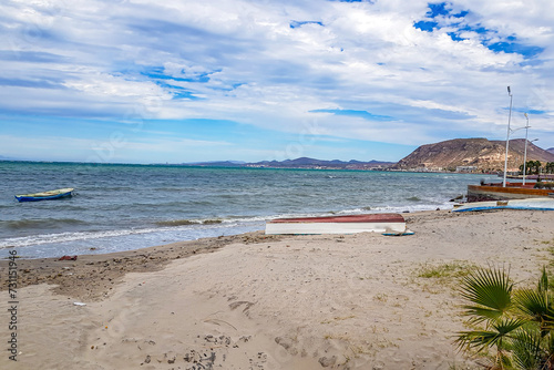 Fototapeta Naklejka Na Ścianę i Meble -  Coastal beach landscape, overturned boat on sand, another in waters in Sea of Cortez, mountains and horizon against blue sky with white clouds in background, day in La Paz, Baja California Sur Mexico