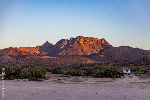 Mexican desert landscape with juniper trees and scrubland, imposing mountain rock formation illuminated by sun in background, car parked on sand, sunset in desert of Baja California Sur, Mexico © Emile