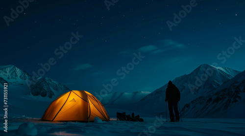 Illuminated yellow tent at night in snowy mountains with person standing next to it and starry sky, long night exposure of tent in mountains