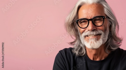 A man with a gray beard and long gray hair wearing glasses smiling and dressed in a black shirt against a pink background.