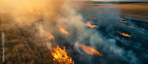 Aerial view of a dry grass meadow field burning during a drought and hot weather, causing a wild open fire that destroys the grass and creates smoke. The incident highlights the problem of air