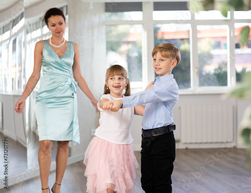 Small passionate ballroom enthusiasts, tween girl and boy practicing graceful waltz routine in well-lit dance hall under female mentor watchful eye..