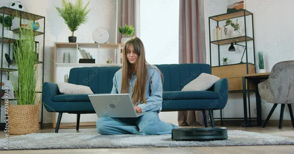 Portrait of woman which resting on carpet at home and listening music on laptop while automatic vacuum cleaner cleaning carpet Portrait of woman which resting on carpet at home and listening music on laptop while automatic vacuum cleaner cleaning carpet