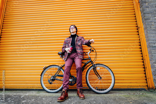 Young smiling hipster male student enjoying coffee out of reusable thermos cup while sitting on cruiser bicycle near orange wall. Eco friendly transport for carbon footprint. Sustainable lifestyle