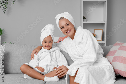 Little girl with her grandmother in bathrobes sitting on sofa at home