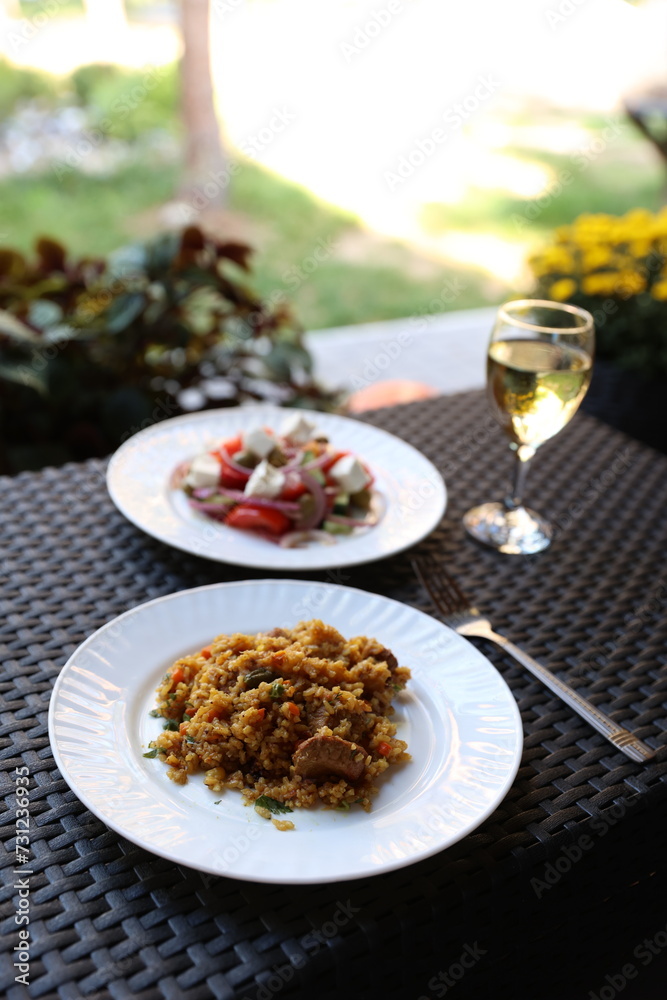 there is food on the rattan table. Pilaf and Greek vegetable salad and a glass of white wine on the table. Eat dinner outside. Garden furniture for home. rattan furniture on the terrace
