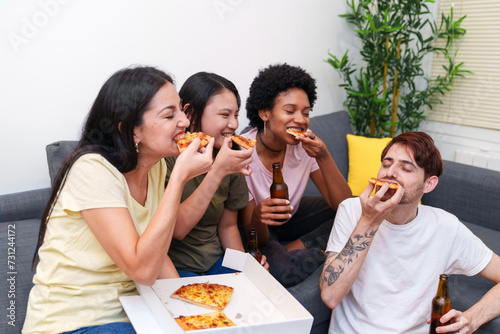 Diverse friends share a joyful moment eating pizza and drinking beer together on a sofa, creating warm memories at a casual get-together