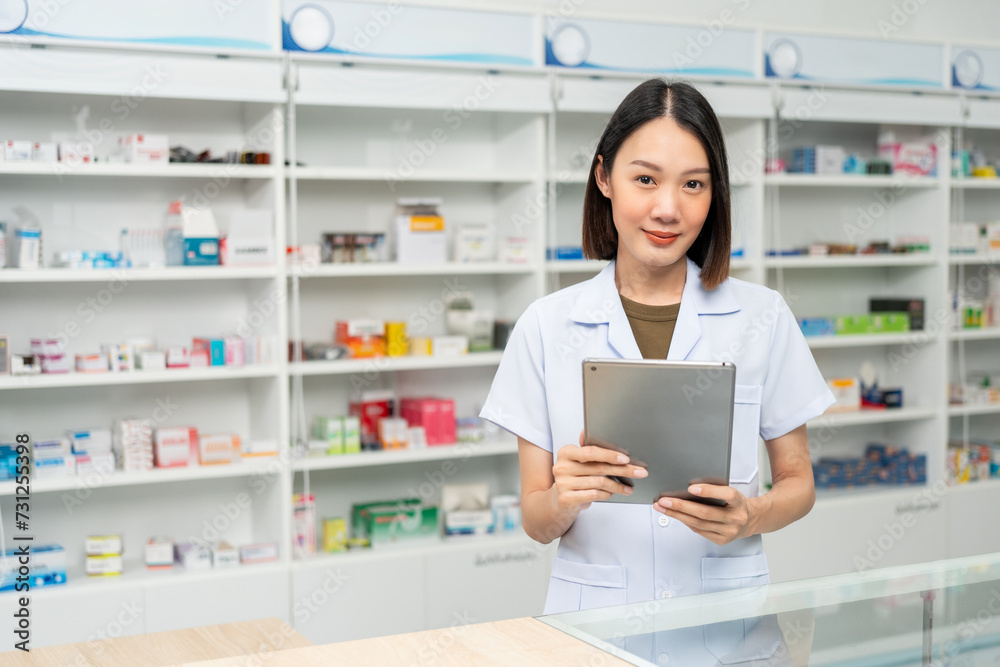Beautiful asian woman pharmacist checks inventory of medicine in pharmacy drugstore. Professional Female Pharmacist wearing uniform standing near drugs shelves working with tablet.