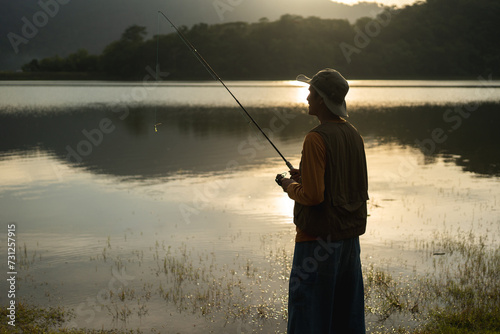 Enjoy moment of Handsome man fishing as a leisure activity during his vacation at the lake on sunset. Silhouette at sunset moment of man fishing rotation with reel. © Chanakon