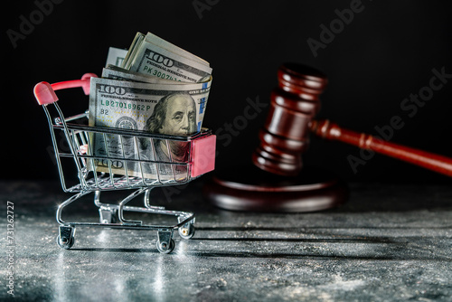 Shopping cart on wheels with dollar bills and wooden judge gavel on dark background, closeup. Finance, business, purchases, spending and sales concept