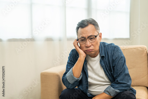 Asian mature old man sitting bored serious thinking in living room at home. Portrait of serious depressed senior asian man. Mature People with problem in life photo