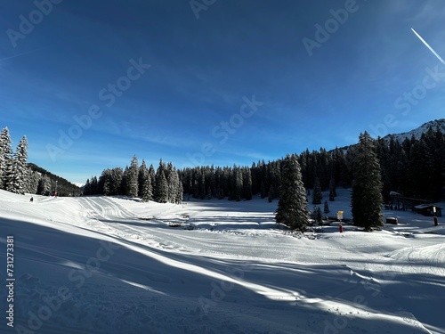 A picturesque mountain slope, blanketed in pristine snow, stretches beneath a clear blue sky. The crisp winter scene invites skiers to carve through the powdery terrain, framed by majestic peaks.