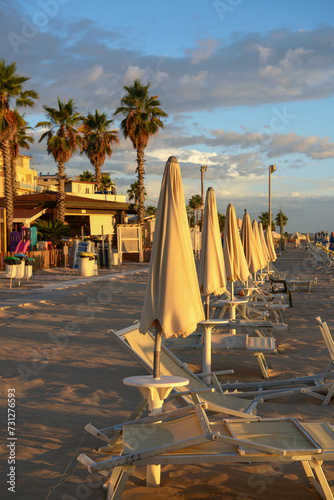 Closed beach umbrellas in the evening on a sandy beach in Italy, sun beds, sand, palm trees and blue sky, holidays with children, relaxation, travel, vacation, background