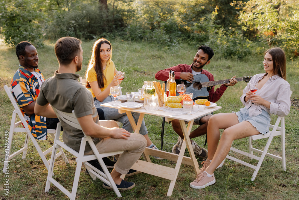Meeting of multiracial group of friends playing guitar, singing, eating dinner and drinking wine during party in the forest Meeting of multiracial group of friends playing guitar, singing, eating dinner and drinking wine during party in the forest