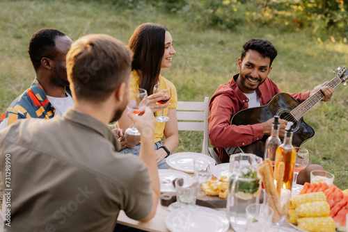 Multiracial group of people, Indian hipster man playing guitar and friends eating dinner and drinking wine during party in the forest