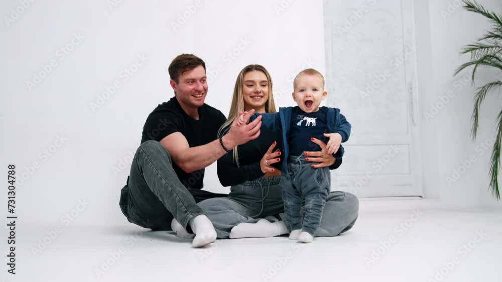 Caucasian parents sit on the floor holding a baby boy. Little kid is cheering and holding hands up. White backdrop. Caucasian parents sit on the floor holding a baby boy. Little kid is cheering and holding hands up. White backdrop.
