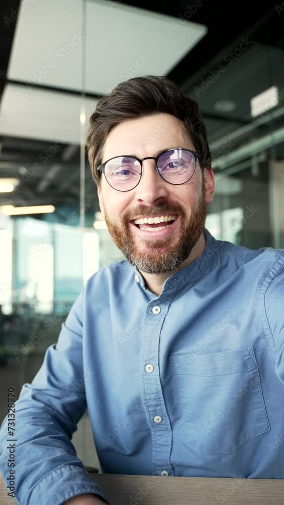 POV Vertical video. Smiling businessman in glasses talking on a video call using a smartphone in a office. Handsome manager waving hand looking at camera. Online chat with a client or business partner POV Vertical video. Smiling businessman in glasses talking on a video call using a smartphone in a office. Handsome manager waving hand looking at camera. Online chat with a client or business partner
