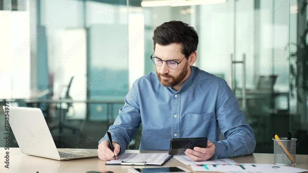 Busy entrepreneur in glasses making financial calculations using calculator, taking notes typing on a laptop sitting at workplace. Confident businessman working fills out documents in business office