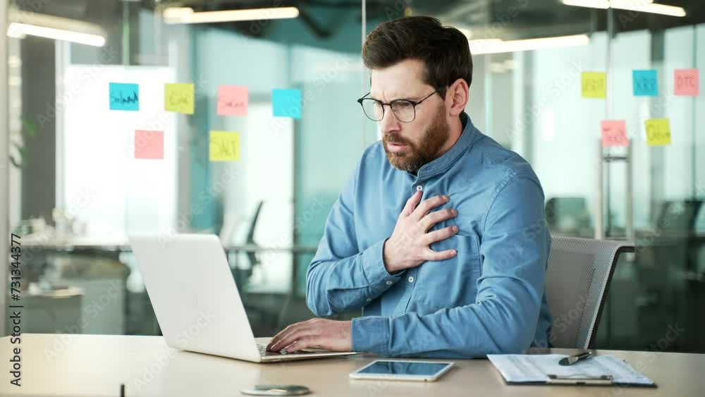 Mature businessman has a heart attack while working on a laptop sitting at a workplace in a business office. The sick employee suffers from tension in the chest and holds the sore spot with his hands Mature businessman has a heart attack while working on a laptop sitting at a workplace in a business office. The sick employee suffers from tension in the chest and holds the sore spot with his hands