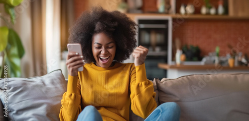 Portrait of young African-American woman celebrating victory, sitting on the couch with a smartphone, raising fist up in triumph, happy black female screams yes photo