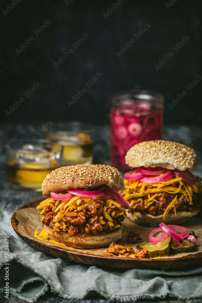 Front shot of Sloppy Joes (minced beef sandwiches) with cheddar cheese, pickled red onions on wood plate with pickle jar in background Front shot of Sloppy Joes (minced beef sandwiches) with cheddar cheese, pickled red onions on wood plate with pickle jar in background