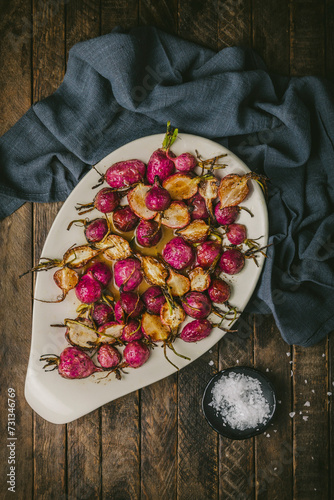 Oval platter with roasted pink radishes in a brown butter sauce on a wood table with a napkin and sea salt photo