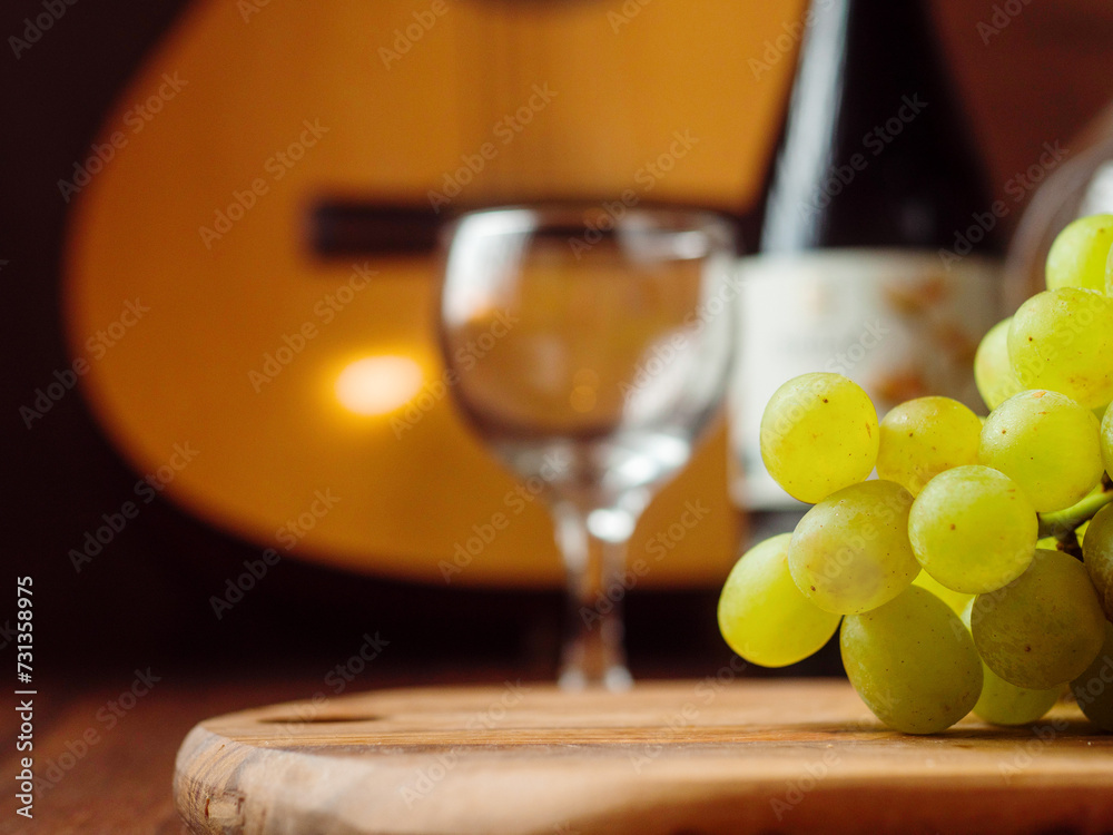 Green grapes in focus, glass and bottle of red wine and classic wooden guitar out of focus in the background.