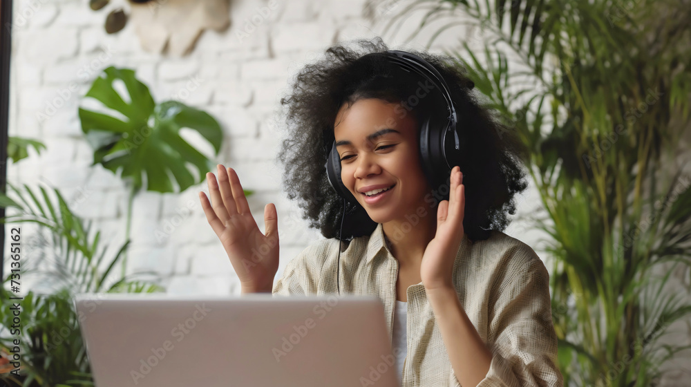 Young African American woman sitting in the living room of a house or home, and using a laptop computer. Female adult freelancing, wearing black headphones or headset, and smiling, customer support Young African American woman sitting in the living room of a house or home, and using a laptop computer. Female adult freelancing, wearing black headphones or headset, and smiling, customer support