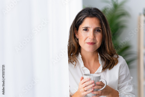 Close-up of a smiling mature woman next to the window drinking coffee