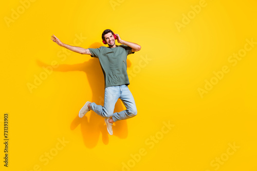Full length photo of cool carefree guy wear khaki t-shirt listen songs headphones jumping high empty space isolated yellow color background