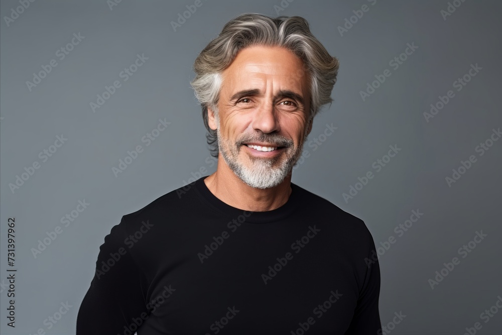 Handsome mature man with grey hair and beard smiling at camera while standing against grey background