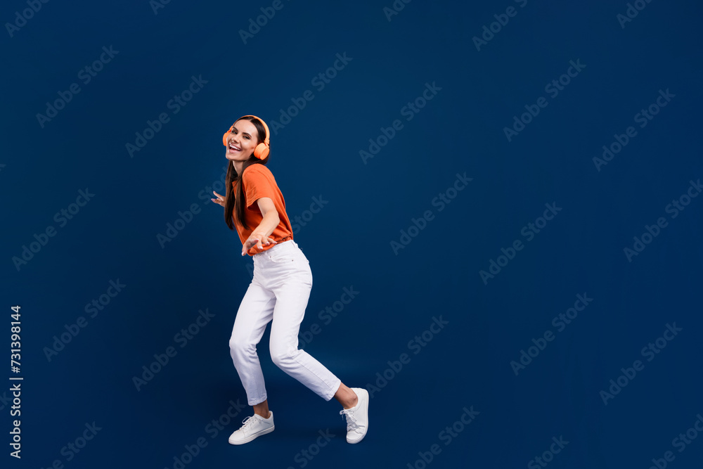 Full length photo of pretty cute lady wear orange t-shirt headphones enjoying discotheque empty space isolated dark blue color background Full length photo of pretty cute lady wear orange t-shirt headphones enjoying discotheque empty space isolated dark blue color background