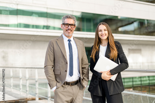 Standing side by side, the seasoned businessman and his young female colleague share a professional moment, woman holding laptops and both dressed in business attire against a modern building backdrop