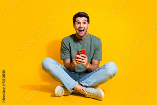 Full length photo of positive impressed guy wear khaki t-shirt communicating modern gadget isolated yellow color background