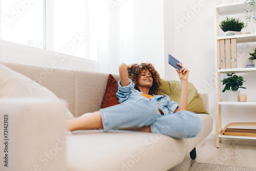 Cheerful woman sitting on a cozy couch at home, holding a mobile phone and smiling while chatting online.