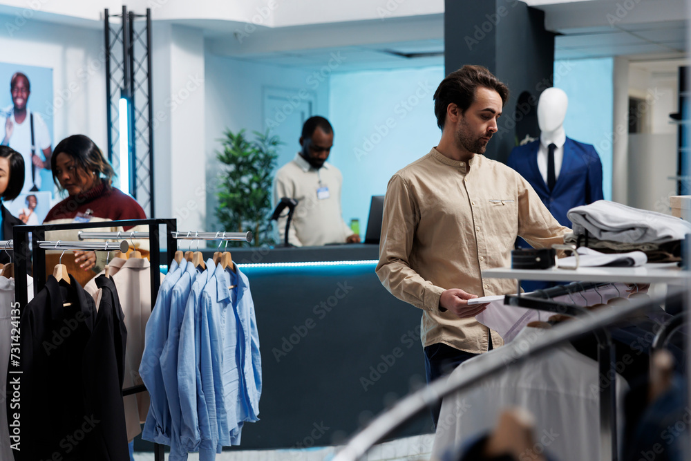 Fototapeta premium Caucasian man browsing through rack of clothing items at store, searching for casual shirt. Fashion boutique customer selecting size while exploring outfit in shopping mall showroom