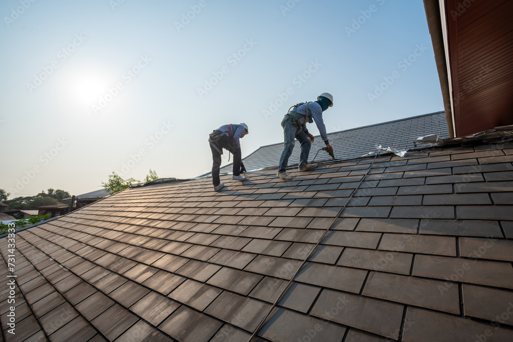 Roofer working in special protective work wear gloves, using air or pneumatic nail gun repair and replace roofing tiles on top of the new roof under construction residential building - obrazy, fototapety, plakaty 