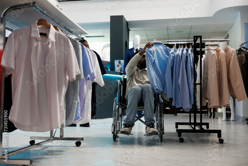 African american man in wheelchair browsing through rack and choosing casual shirt in clothing shop. Shopping center boutique customer with physical disability examining apparel