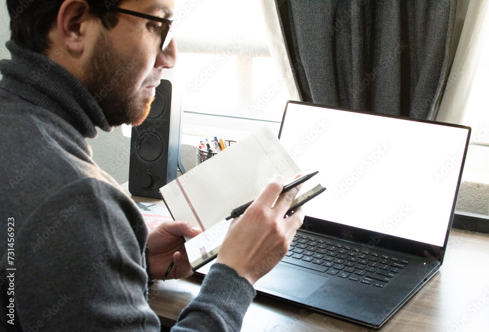 Joven apuesto con gafas de bella sonrisa y suéter gris sosteniendo un cuaderno y trabajando en el portátil desde su hogar. Trabajo en casa Joven apuesto con gafas de bella sonrisa y suéter gris sosteniendo un cuaderno y trabajando en el portátil desde su hogar. Trabajo en casa