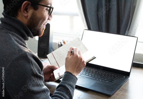 Joven apuesto con gafas de bella sonrisa y suéter gris sosteniendo un cuaderno y trabajando en el portátil desde su hogar. Trabajo en casa photo