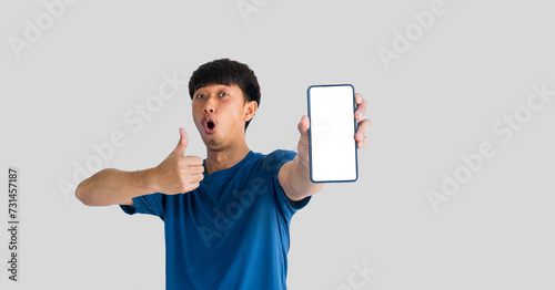 Asian man in blue t-shirt makes wow face and gives thumbs up on mobile phone with blank white smartphone screen isolated on gray background. People recommending new applications or mobile websites