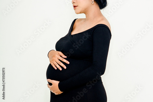 Pregnant woman in elegant dres holds hands on belly on a white background at studio. Pregnancy, maternity, preparation concept. Close-up, copy space, indoors.