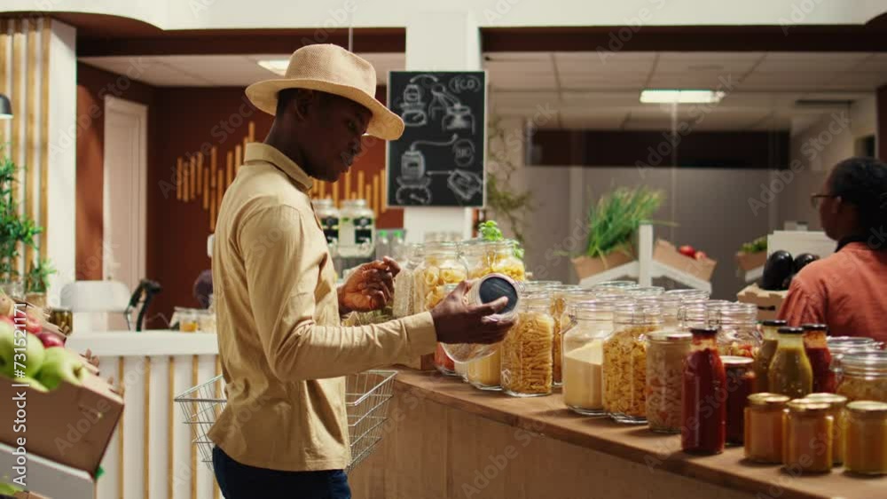 Client examines cereals and grains at zero waste eco store, looking for natural organic products and pantry supplies. African american man buying homemade spices and pasta in jars. Camera B. Client examines cereals and grains at zero waste eco store, looking for natural organic products and pantry supplies. African american man buying homemade spices and pasta in jars. Camera B.